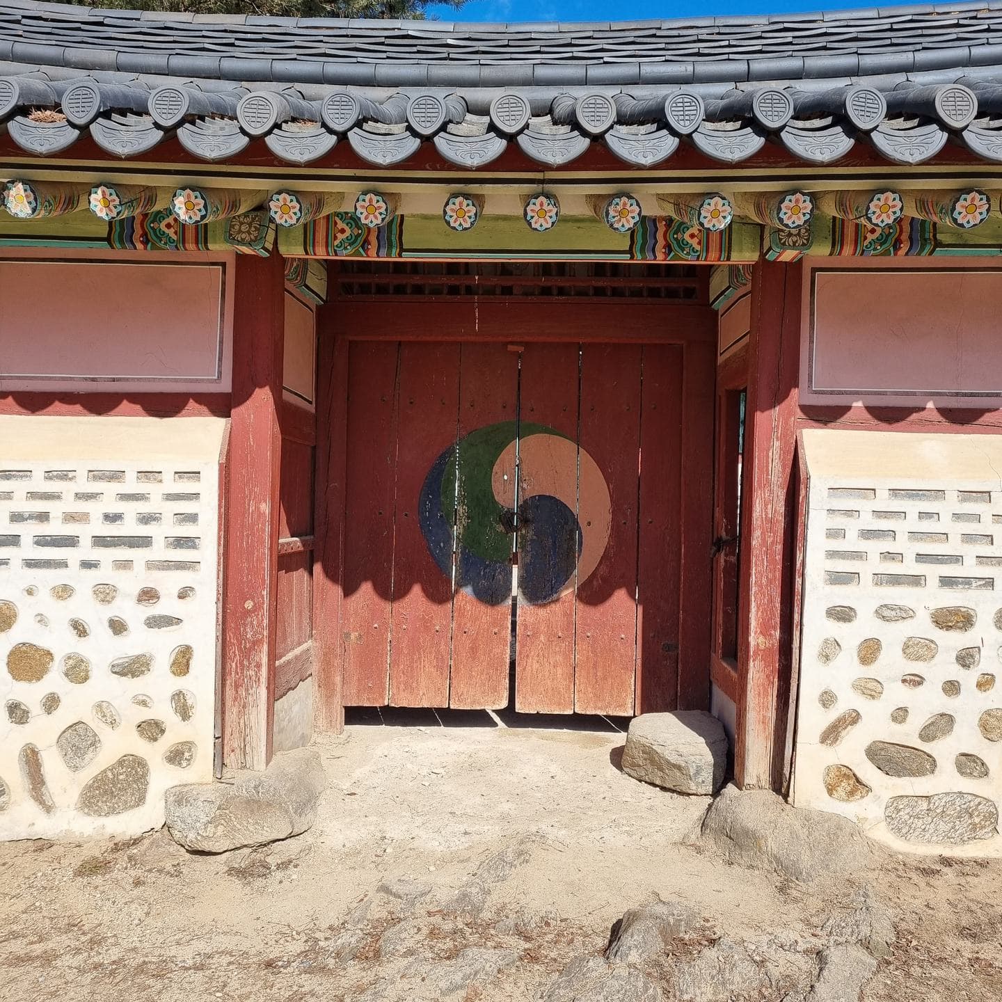 A red wooden gate with a taegeuk symbol, set in a stone and plaster wall