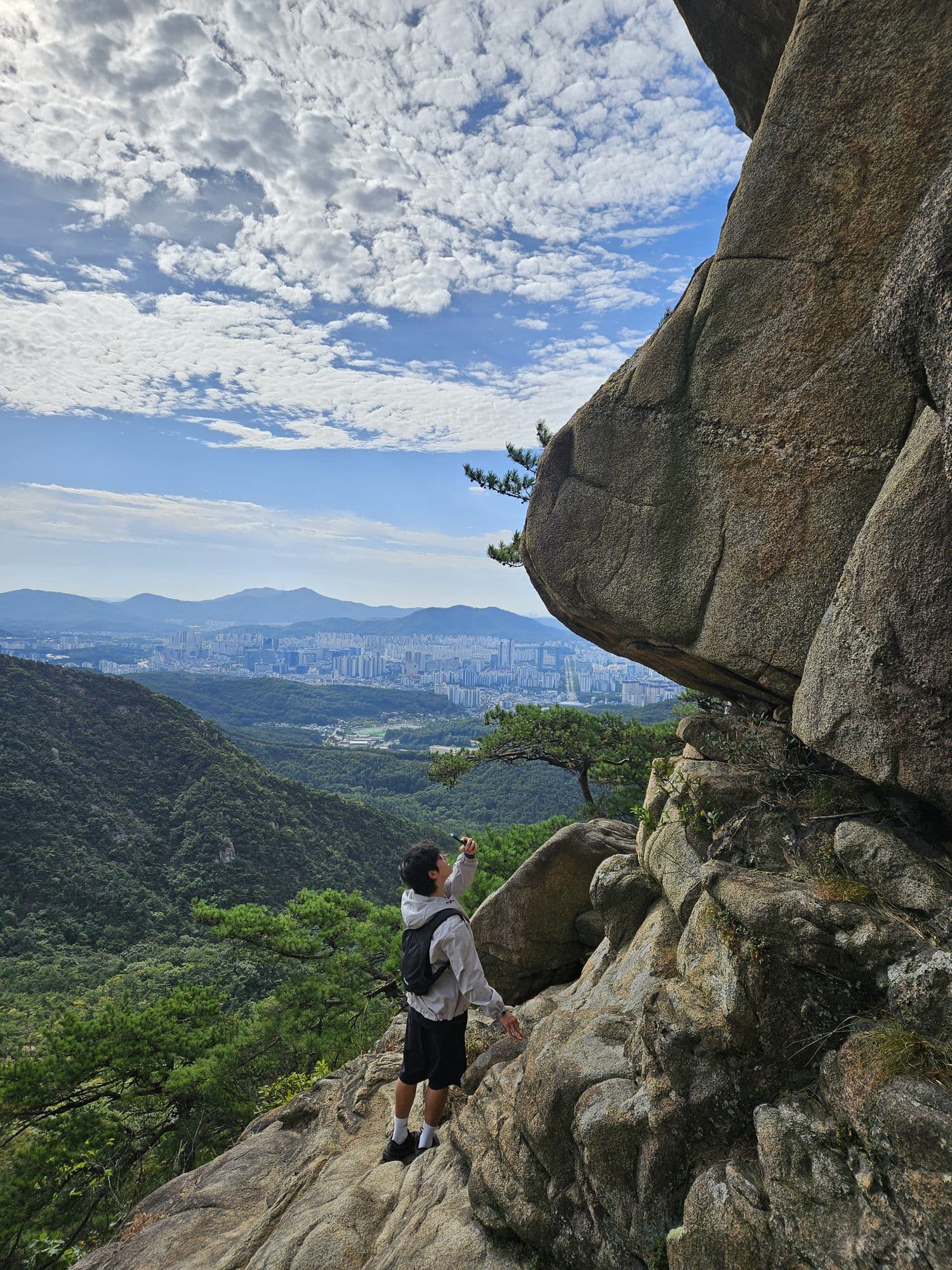 Standing under a huge granite overhang, phone raised, the valley and city far below
