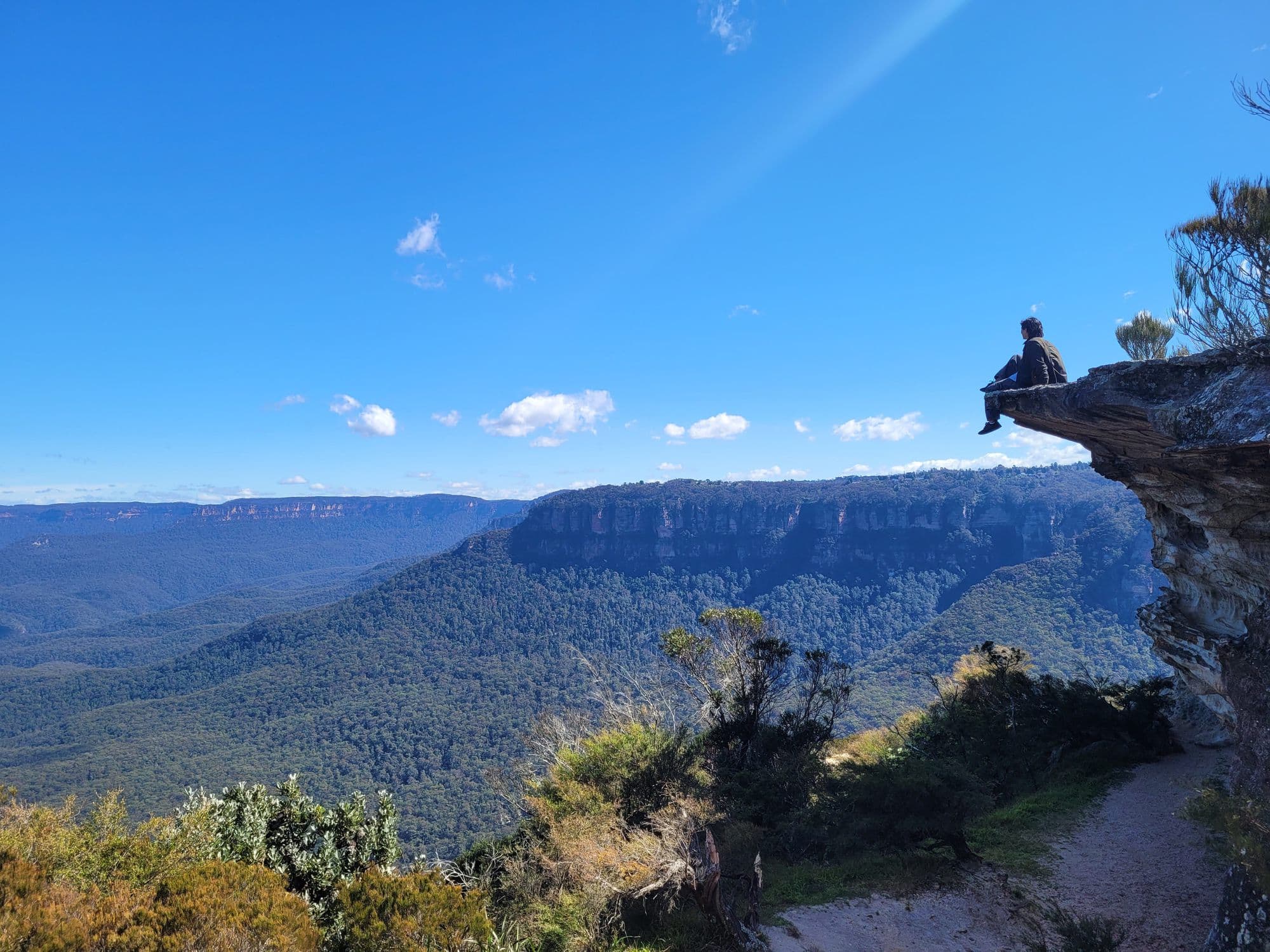 A figure sitting on a sandstone ledge high above a forested valley