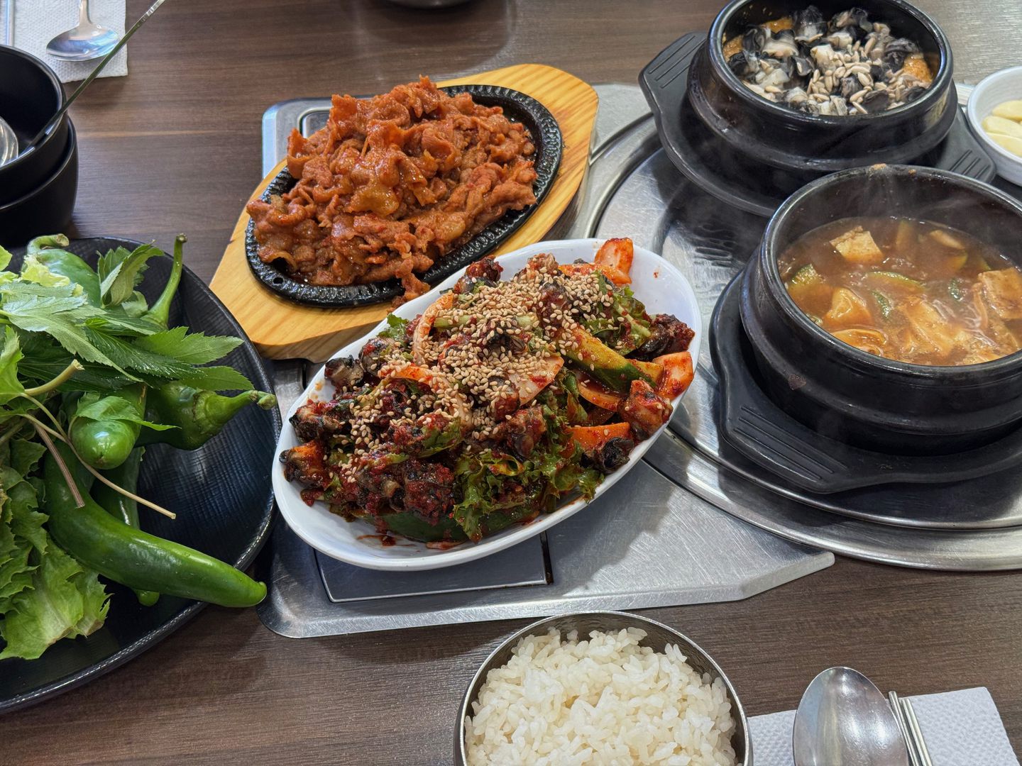 소쿠리에 담긴 상추와 쌈 채소, 풋고추 옆에 제육, 불고기, 뚝배기와 공깃밥이 놓인 모습