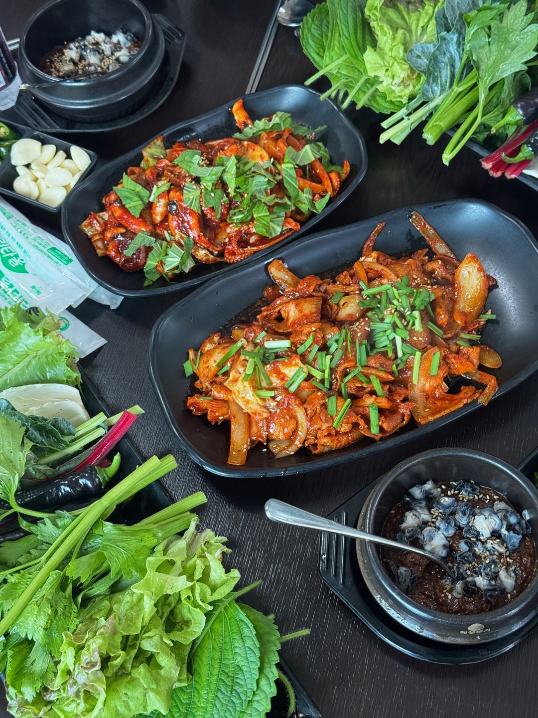 검은 접시 두 개에 담긴 쭈꾸미볶음, 쌈 채소, 마늘과 우렁 강된장 뚝배기가 차려진 테이블을 위에서 찍은 사진