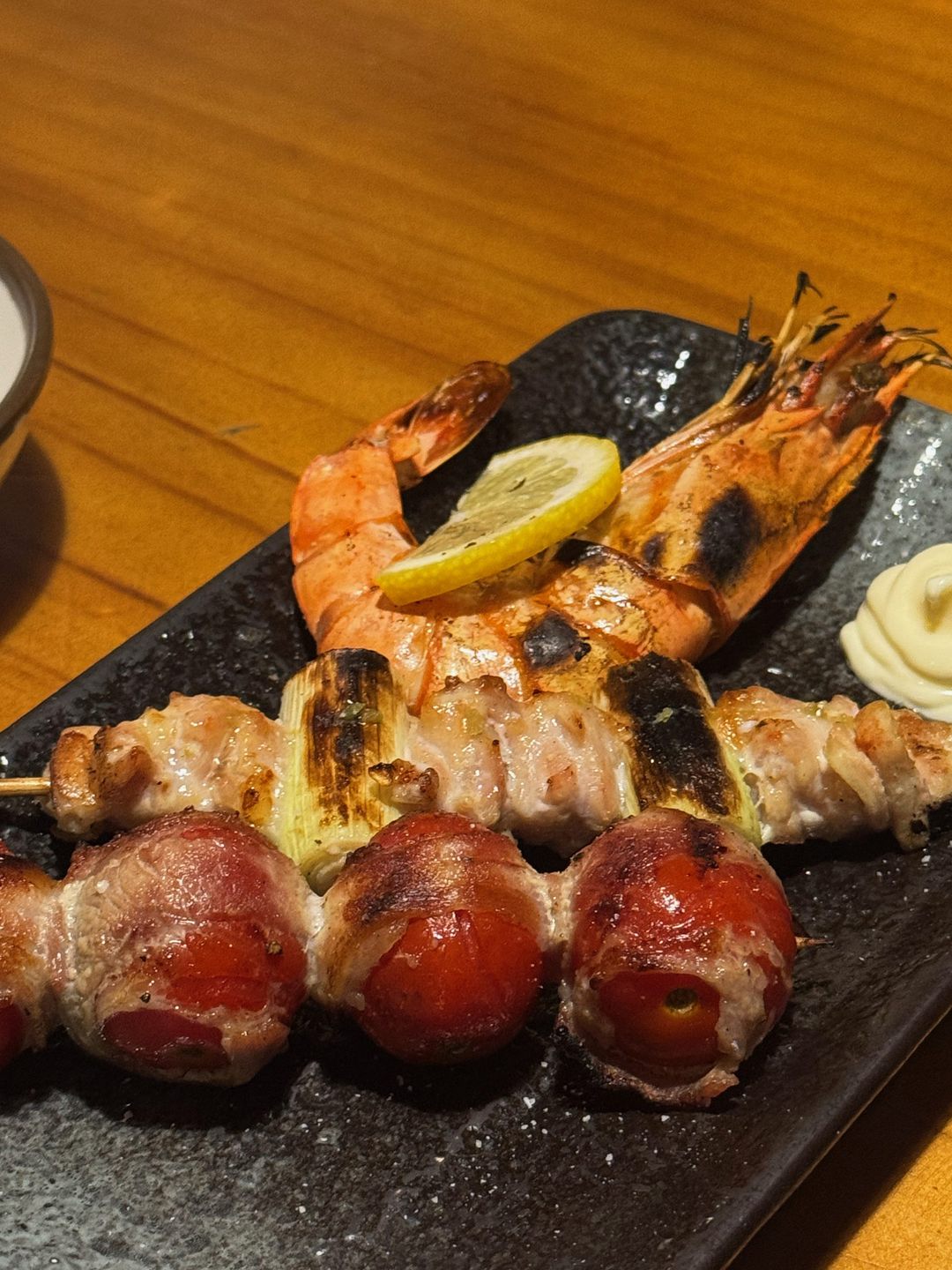 검은 접시 위의 통새우 꼬치, 대파 고기 꼬치, 방울토마토 베이컨말이 꼬치와 레몬