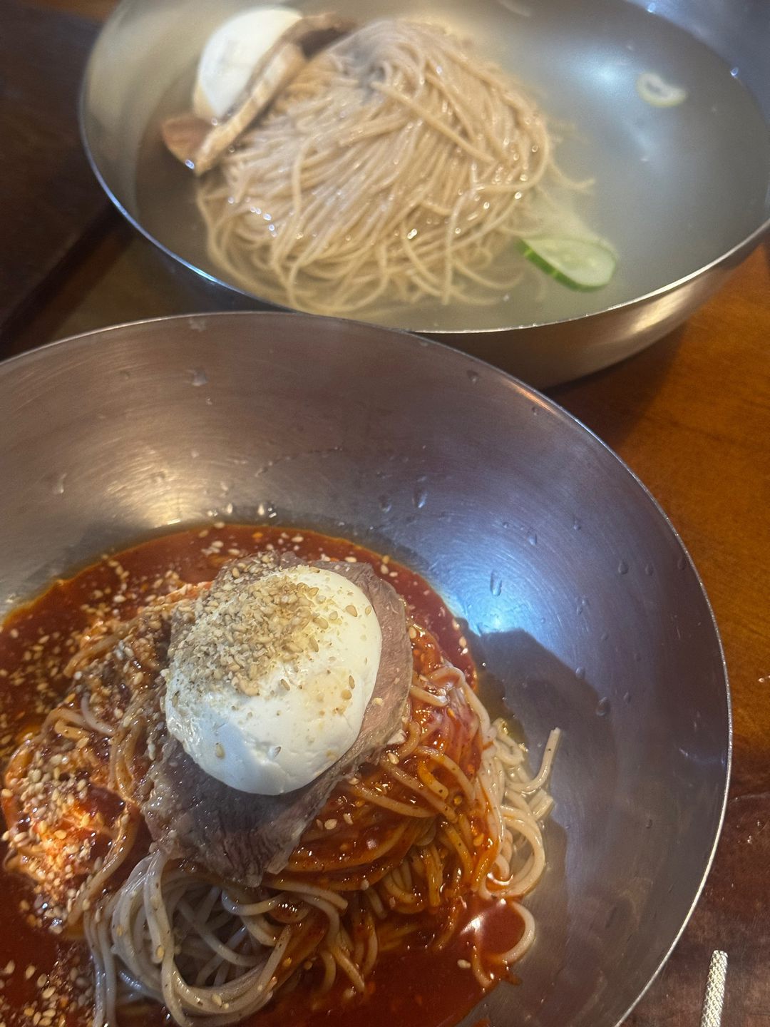 빨간 양념 위에 반숙 계란과 깨를 올린 비빔냉면, 뒤로 평양냉면 그릇이 보인다