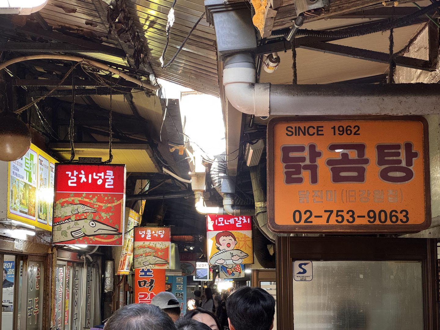 남대문시장 좁은 골목에 걸린 오렌지색 닭곰탕 간판, SINCE 1962와 닭진미(구 강원집), 전화번호가 적혀 있다
