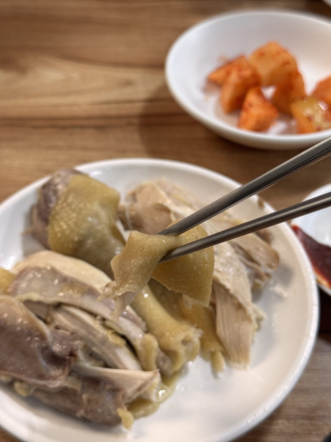 젓가락으로 집어 든 닭 껍질 한 점, 뒤로 깍두기 접시가 보인다